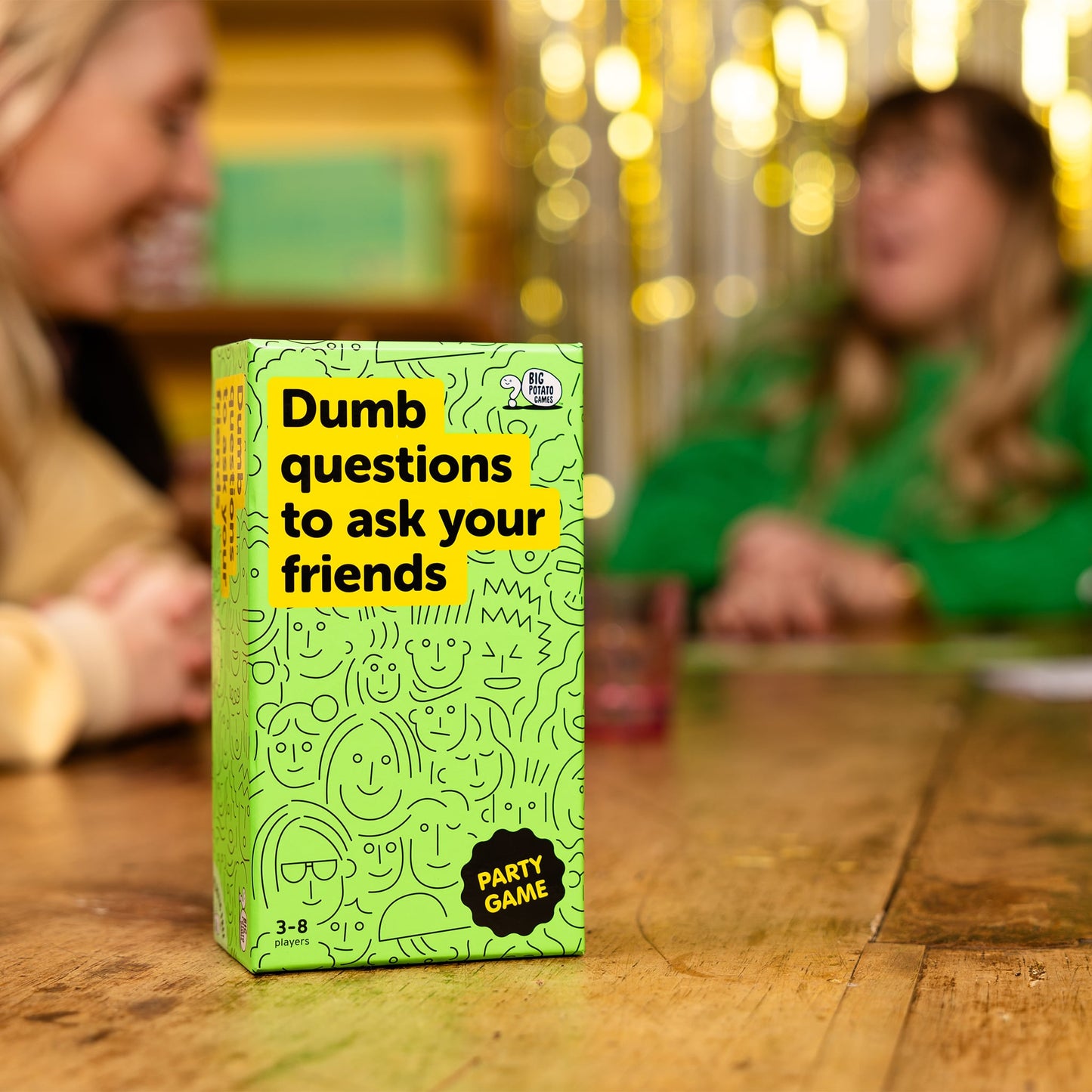 Green game box titled 'Dumb questions to ask your friends' on a wooden table with blurred people in the background.