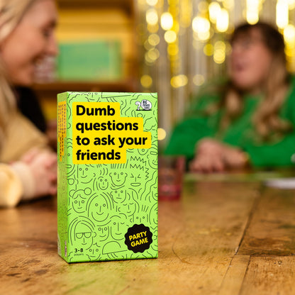 Green game box titled 'Dumb questions to ask your friends' on a wooden table with blurred people in the background.