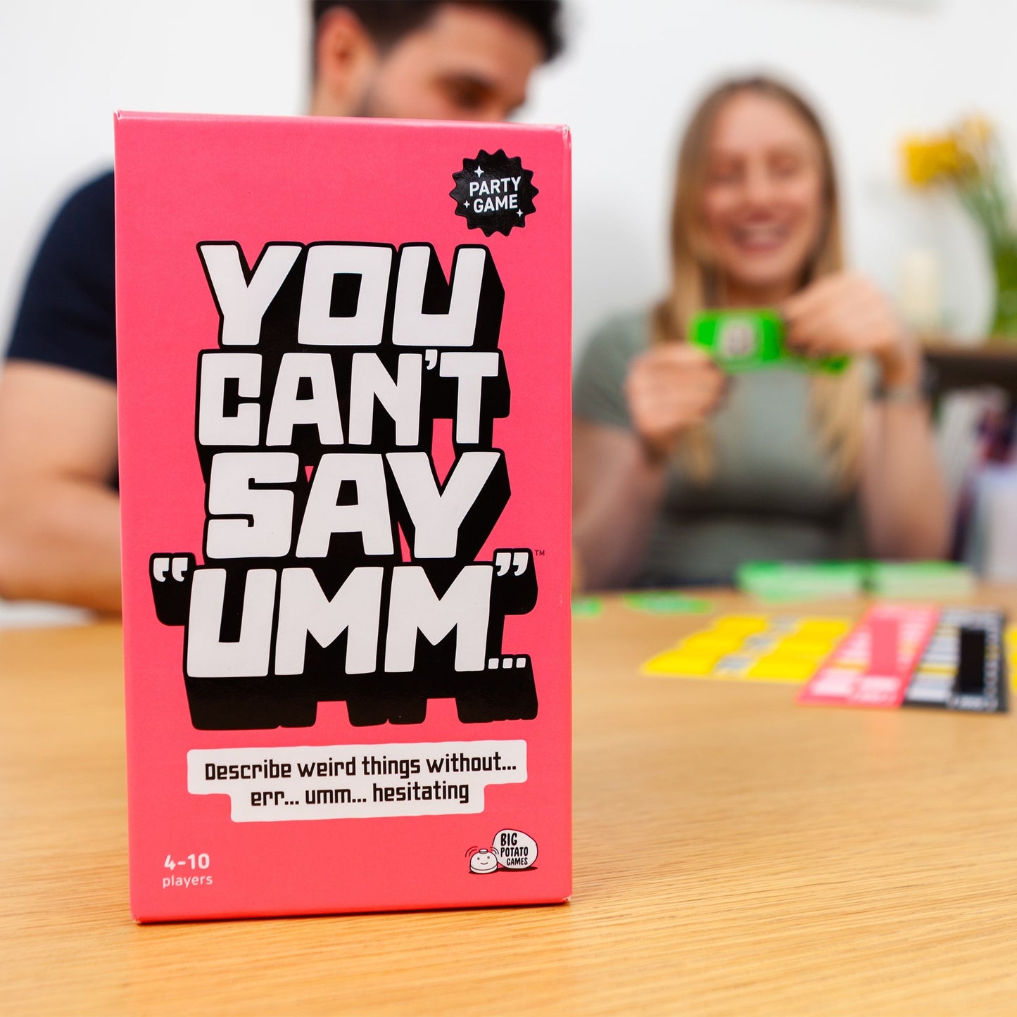 Pink game box titled 'You Can't Say Umm' on a table with people playing and laughing in the background.