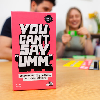 Pink game box titled 'You Can't Say Umm' on a table with people playing and laughing in the background.