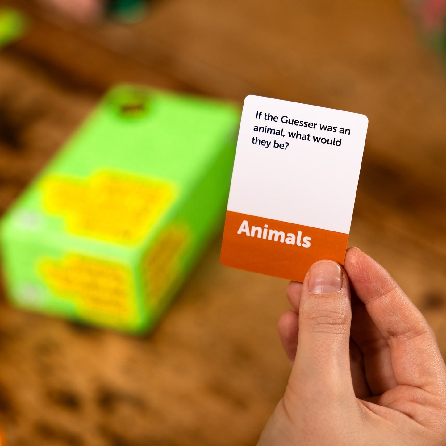 Hand holding a card from the game Dumb Questions To Ask Your Friends with text 'If the Guesser was an animal, what would they be?' and 'Animals' on a blurred background.