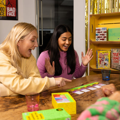 Two women playing the board game Dumb Questions To Ask Your Friends together at a table with books and snacks in the background.